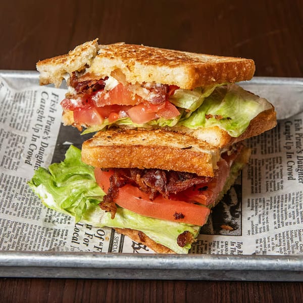 A Port City Cheesesteak BLT sandwich, cut in half, on a metal tray lined with newspaper.