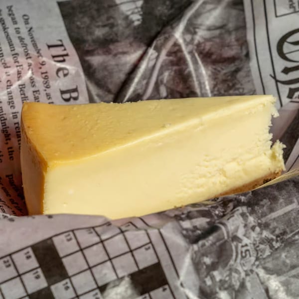 A slice of cheesecake with a golden-brown top, resting on newspaper-patterned wrapping paper.