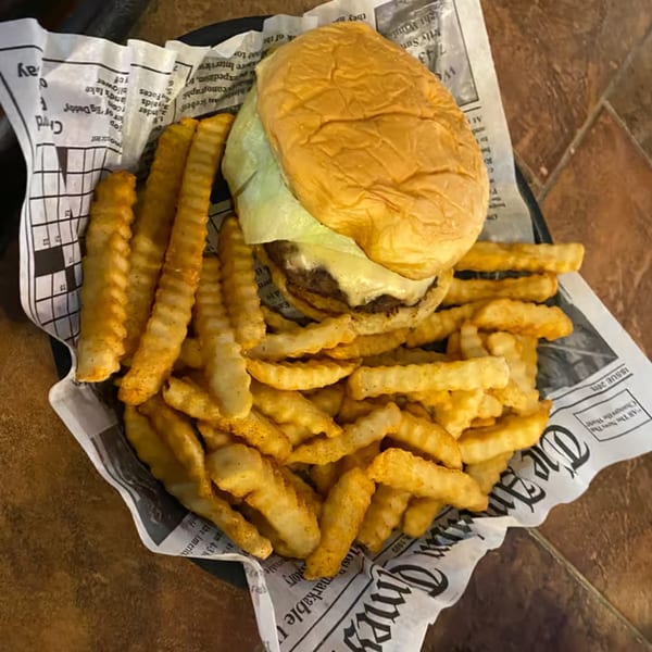 A Port City Cheesesteak burger with melted cheese and lettuce, served with crinkle-cut fries on newspaper.
