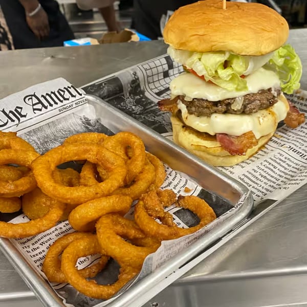 Port City Cheesesteak's burger with bacon, cheese, lettuce, and tomato, served with onion rings.