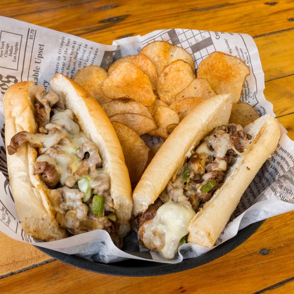 Two Port City Cheesesteak sandwiches with melted cheese and green peppers, served with potato chips.