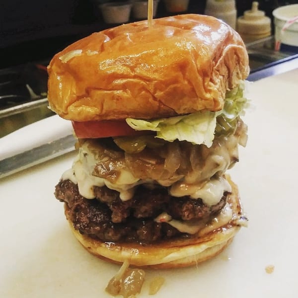A towering Port City Cheesesteak burger with double patties, cheese, onions, lettuce, and tomato.