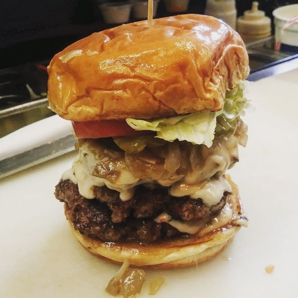 A towering Port City Cheesesteak burger with double patties, cheese, onions, lettuce, and tomato.